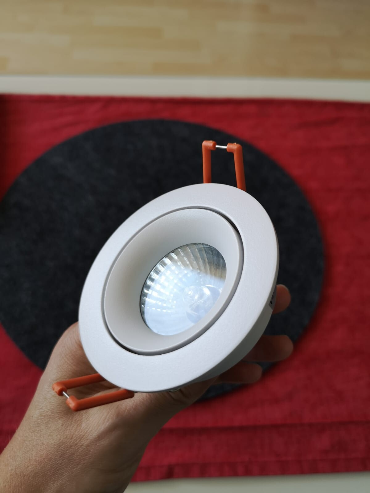 Hand hält runden weißen LED-Deckenstrahler mit orangefarbenen Halterungen über rotem Teppich.