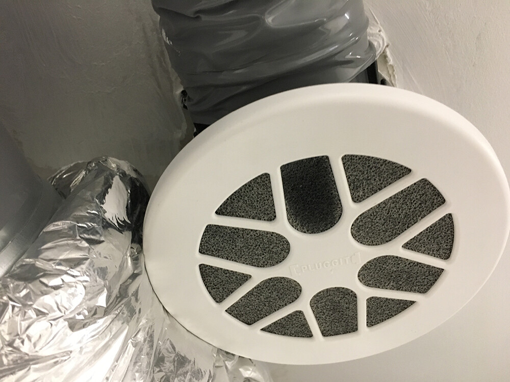 Rundes Lüftungsgitter mit schwarzen Filtern, umgeben von silbernen Flexschläuchen.