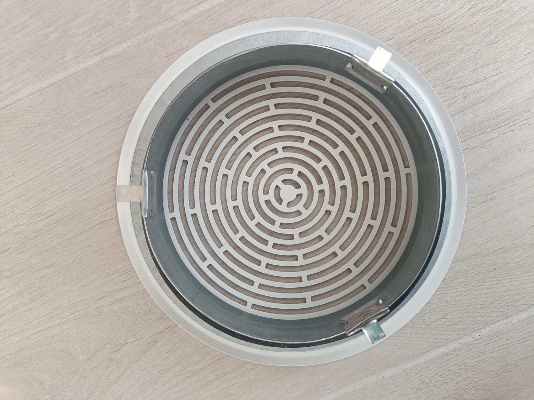 Runder Metalldeckel mit labyrinthartigem Gittermuster und zwei Verriegelungen auf Holzboden