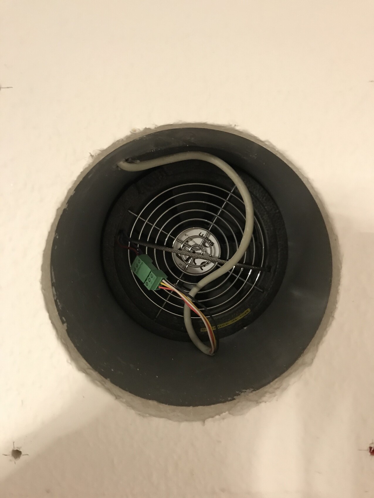 Runder Deckenlüfter hinter Ventilationsgitter, sichtbare Drahtkabel und grüne Klemmleiste.