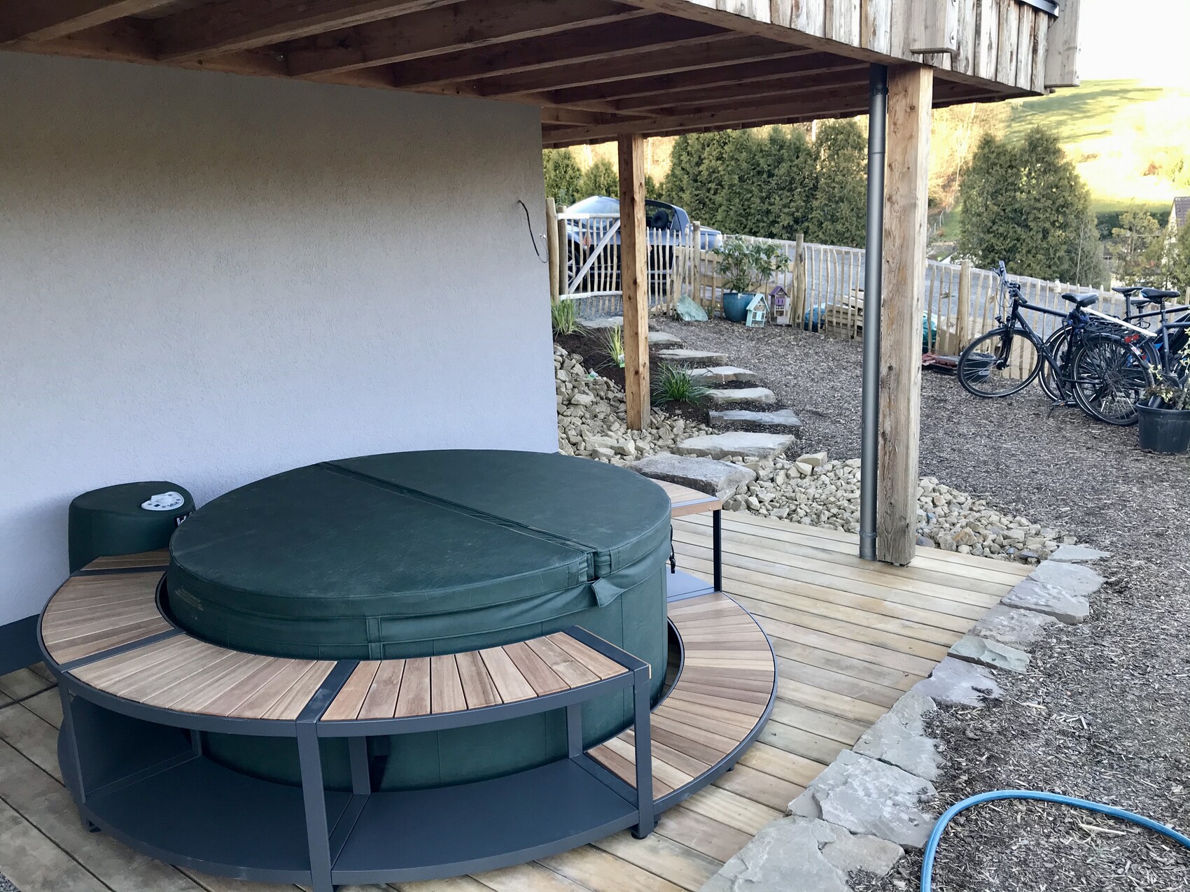 Großer runder Outdoor-Whirlpool mit Abdeckung auf Holzdielen; Garten und Fahrräder im Hintergrund.