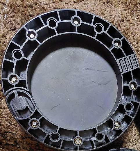 Runder schwarzer Kunststoffdeckel mit Bolzenring, liegt auf braunem Teppich.