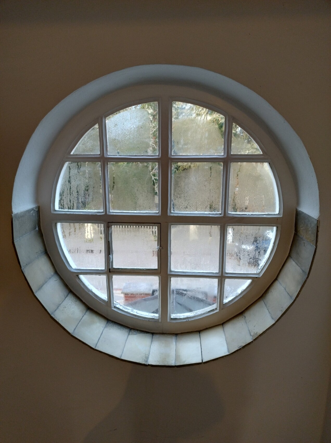 Rundes Fenster mit 4x4-Gitter aus Glas, Blick nach außen auf Bäume und Autos.