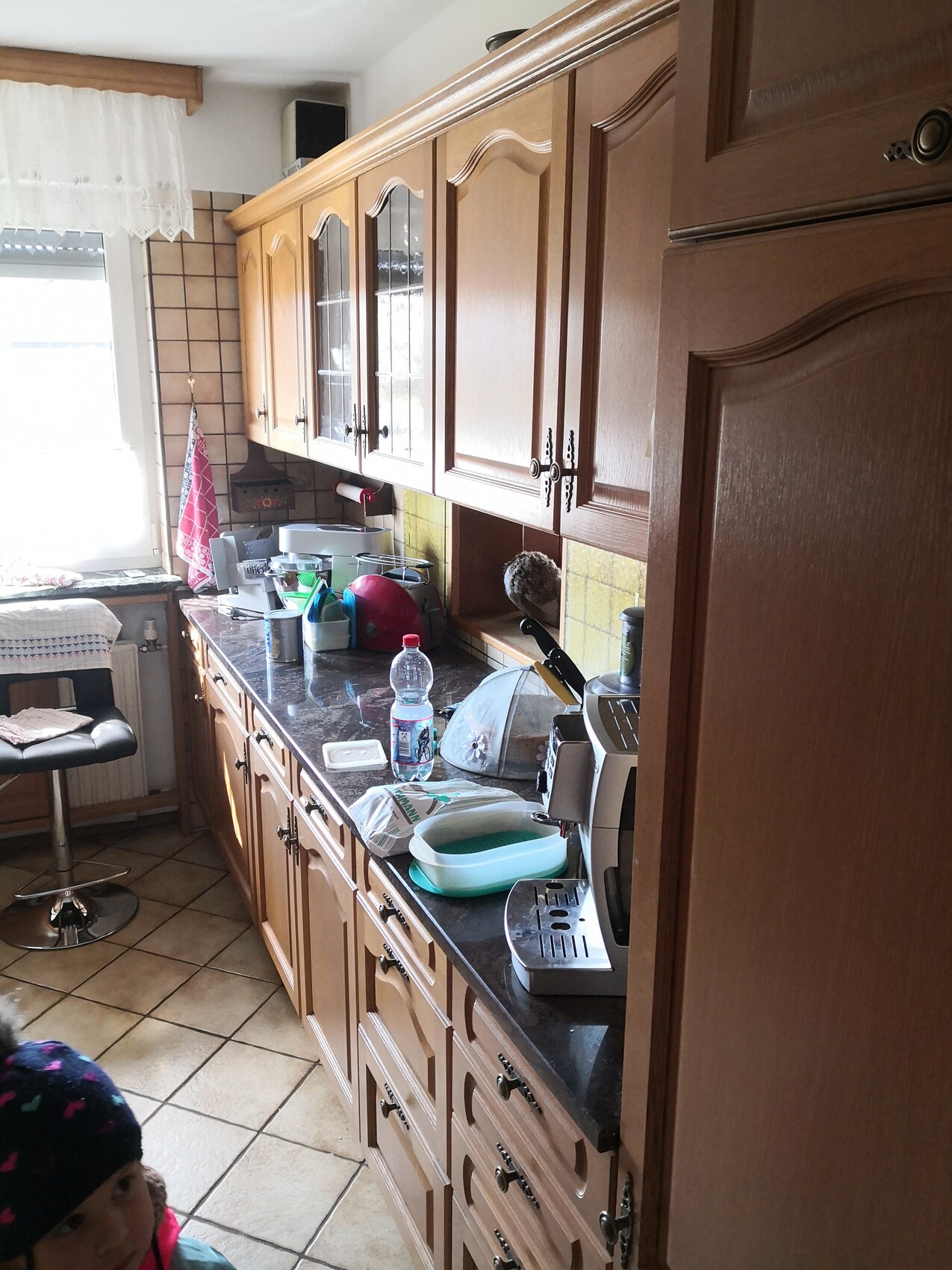 Küche mit Holzmöbeln, volle Arbeitsplatte, Geschirr, Wasserflasche und Toaster; Kind unten links.