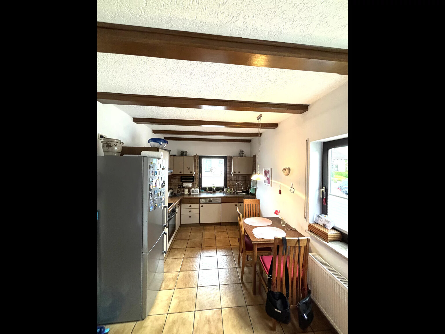 Küche mit Holzbalkendecke, Kühlschrank links, Esstisch rechts nahe Fenster, Fliesenboden.