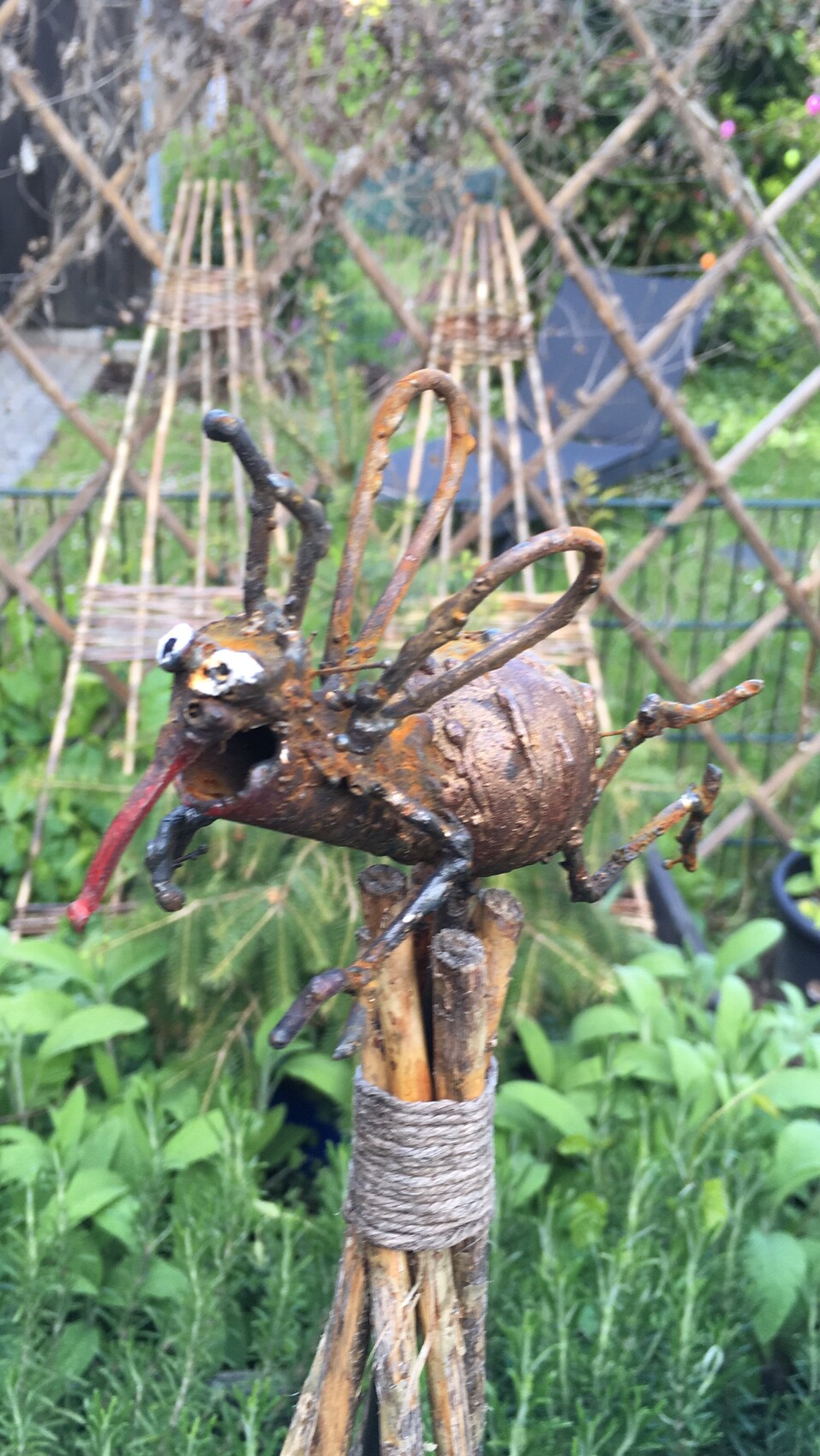 Rostige Metallskulptur eines tierähnlichen Wesens mit roter Zunge, auf Stäben gebunden, im Garten.