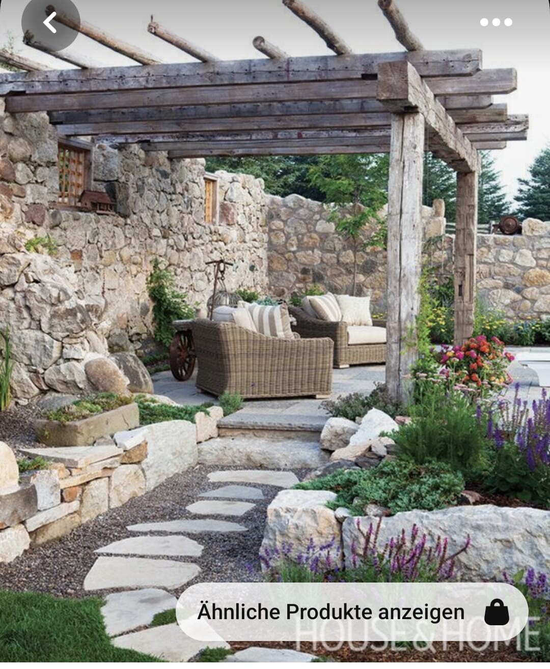 Rustikale Steinmauer mit Holz-Pergola und bequemen Korbmöbel-Sitzgruppen im Garten.
