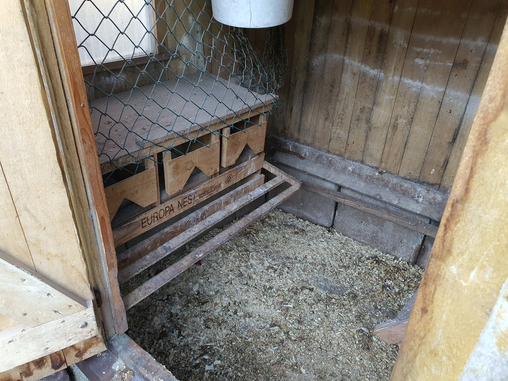 Innenansicht eines Hühnerstalls: Holzwände, Drahtgitter, Nistkästen mit Einstreu am Boden.