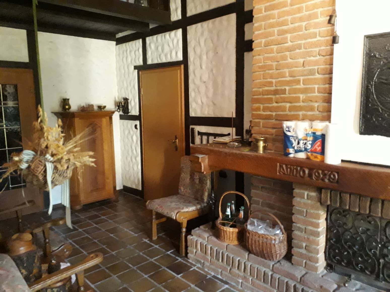Rustikales Wohnzimmer mit Kamin, Holzbalken, brauner Tür, gepolsterter Stuhl und Körben.