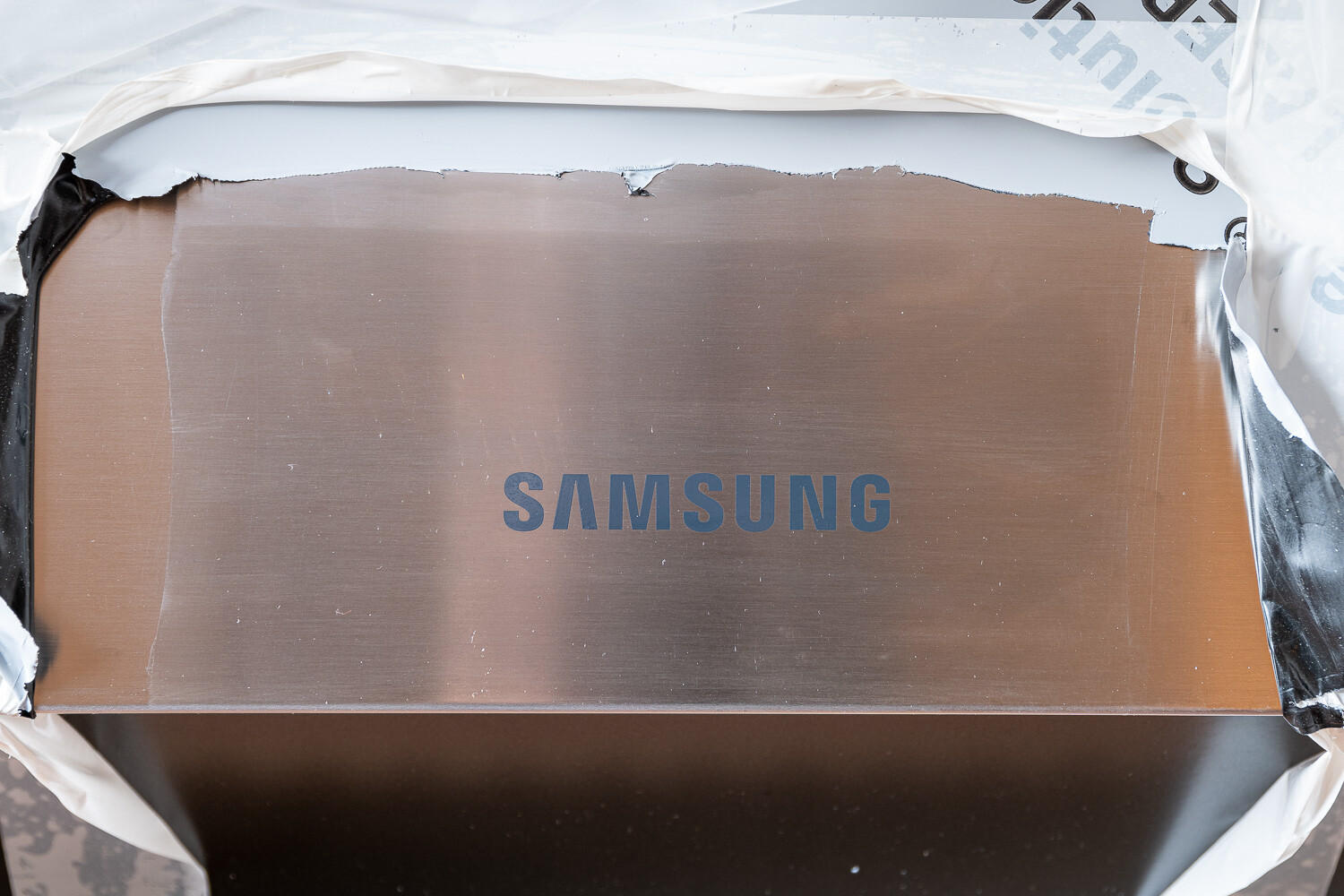 SAMSUNG-Logo auf einer bräunlich-metallischen Oberfläche, von weißer Schutzfolie umgeben.