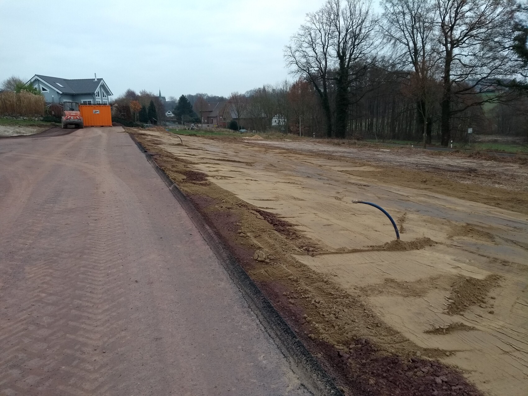 Baustelle an Wohnstraße: Sandboden, blaues Rohr, orange Baucontainer im Hintergrund.