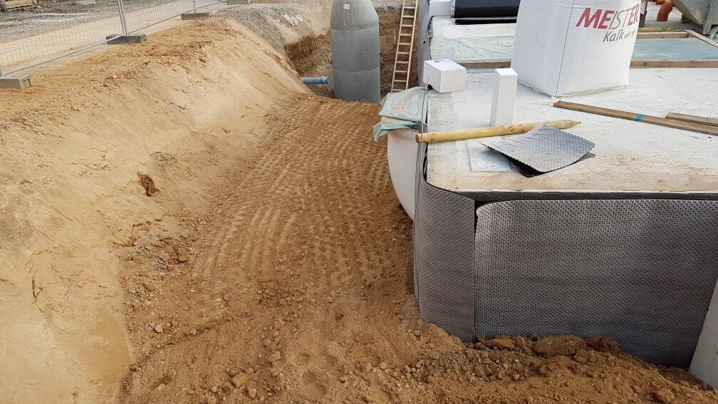 Baugrube mit Sand neben einem metallverkleideten Fundamentkasten; Schaufel liegt darauf.