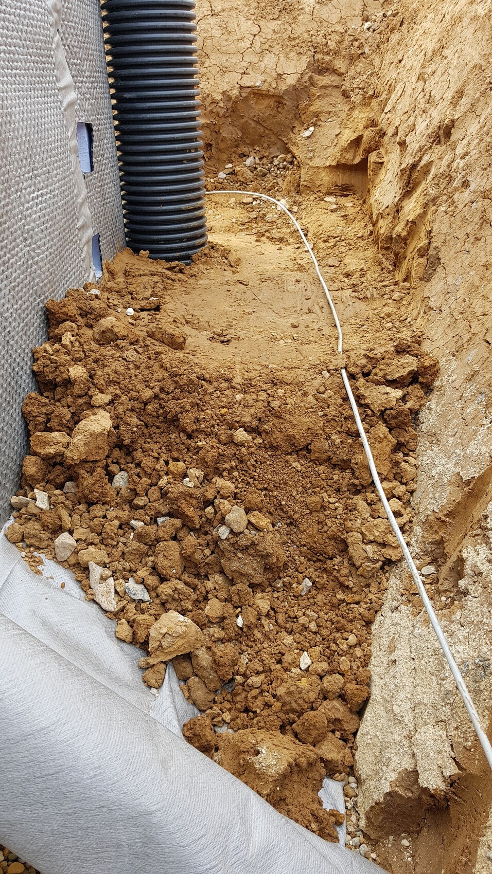 Graben im Baugrund mit schwarzem Rohr und weißer Leitung.