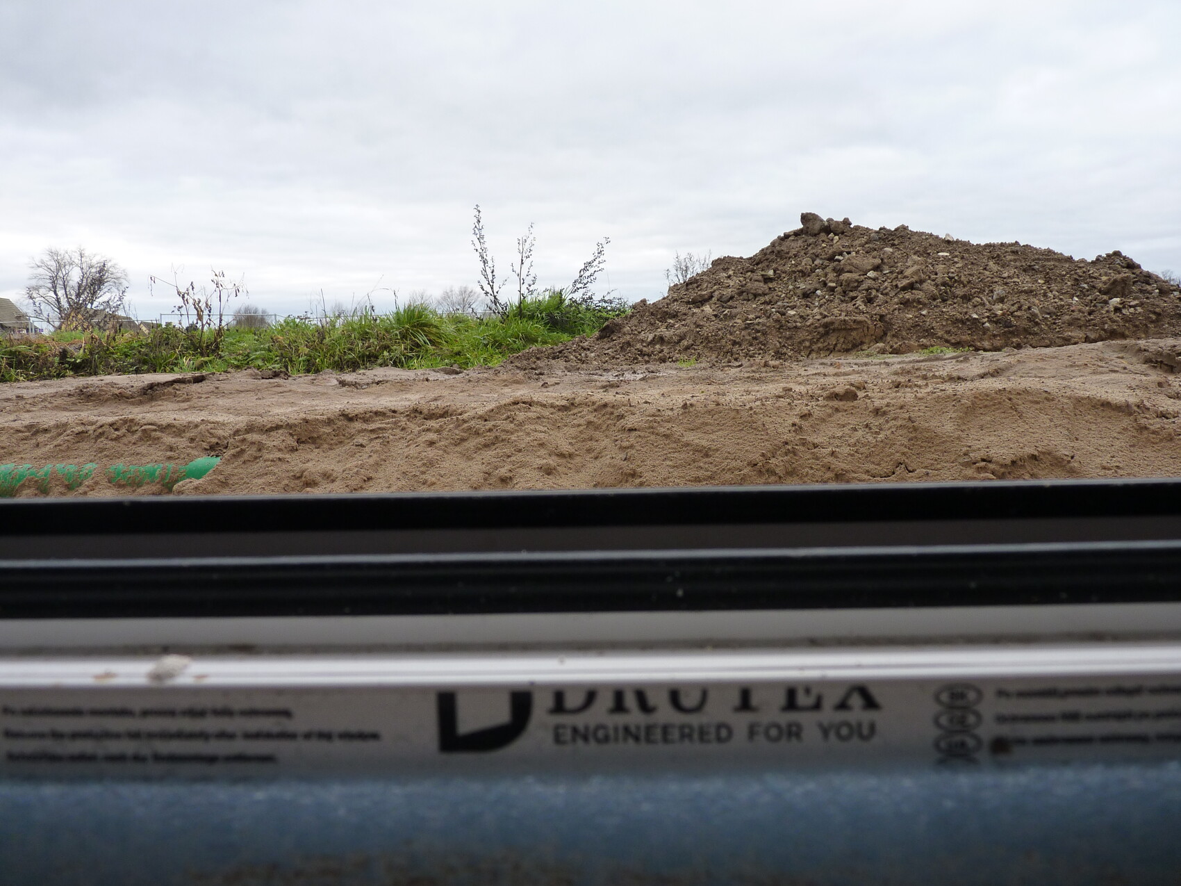 Blick aus einem Fahrzeugfenster auf einen Erdhaufen vor Baugelände mit Gras am Rand und Wolkenhimmel