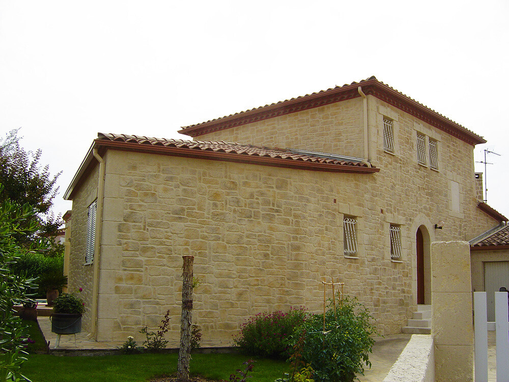 Zweistöckiges Natursteinhaus mit rotem Ziegeldach, bogenförmigem Eingang und Garten davor.