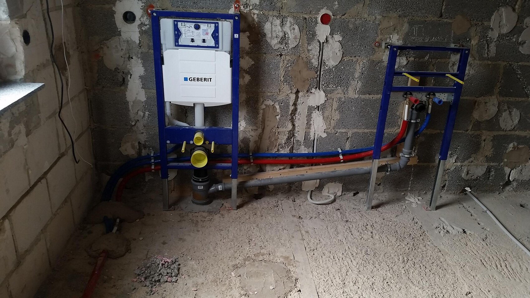 Installationsrahmen mit Geberit-Spülkasten an Wand, sichtbare blaue und rote Wasserleitungen.
