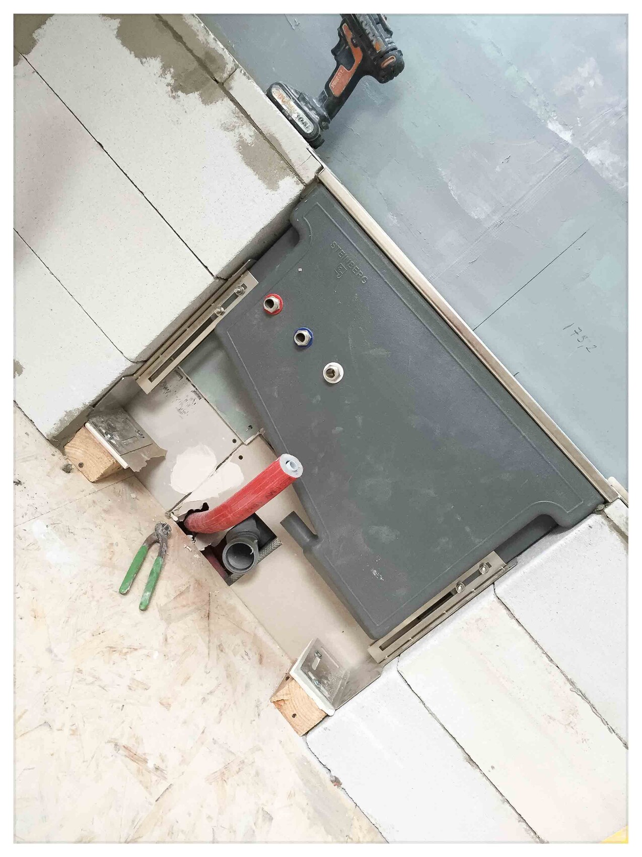 Baustelle: graues Installationspanel mit rotem Flexrohr und grüner Zange am Boden.