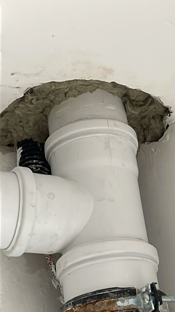 Weißes PVC-Rohr mit T-Verbindung durch Loch in der Wand; daneben flexibles schwarzes Rohr.