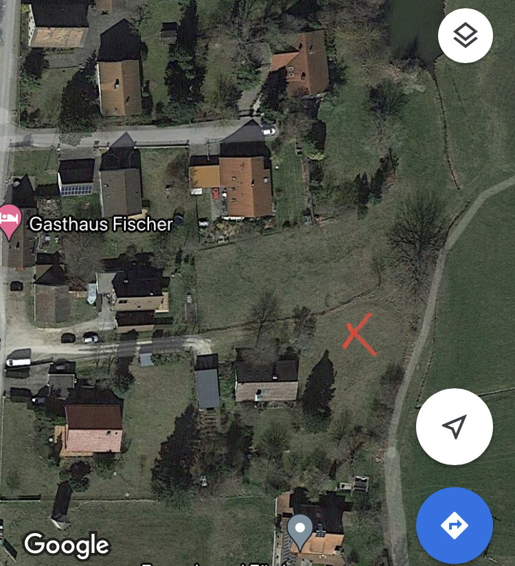 Satellitenbild eines Dorfes mit Häusern, Straßen und roter X-Markierung auf freier Wiese.