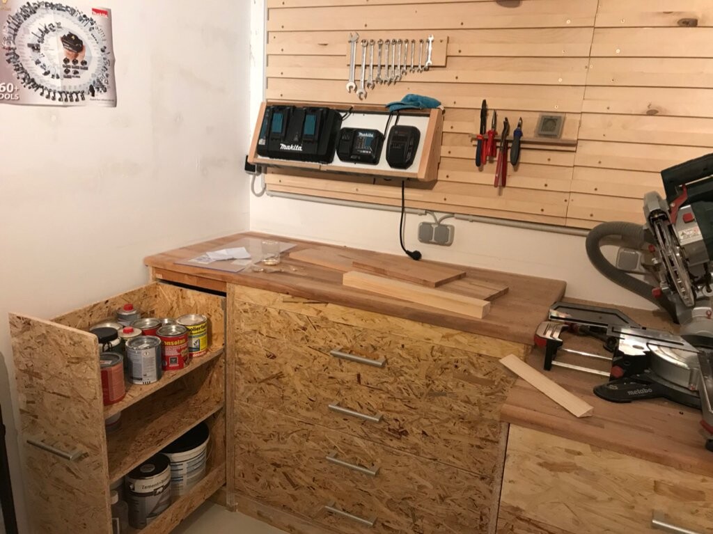 Holzwerkstatt-Eckbereich: Arbeitsbank, OSB-Schränke, Farbdosen, Wandwerkzeug und eine Säge.