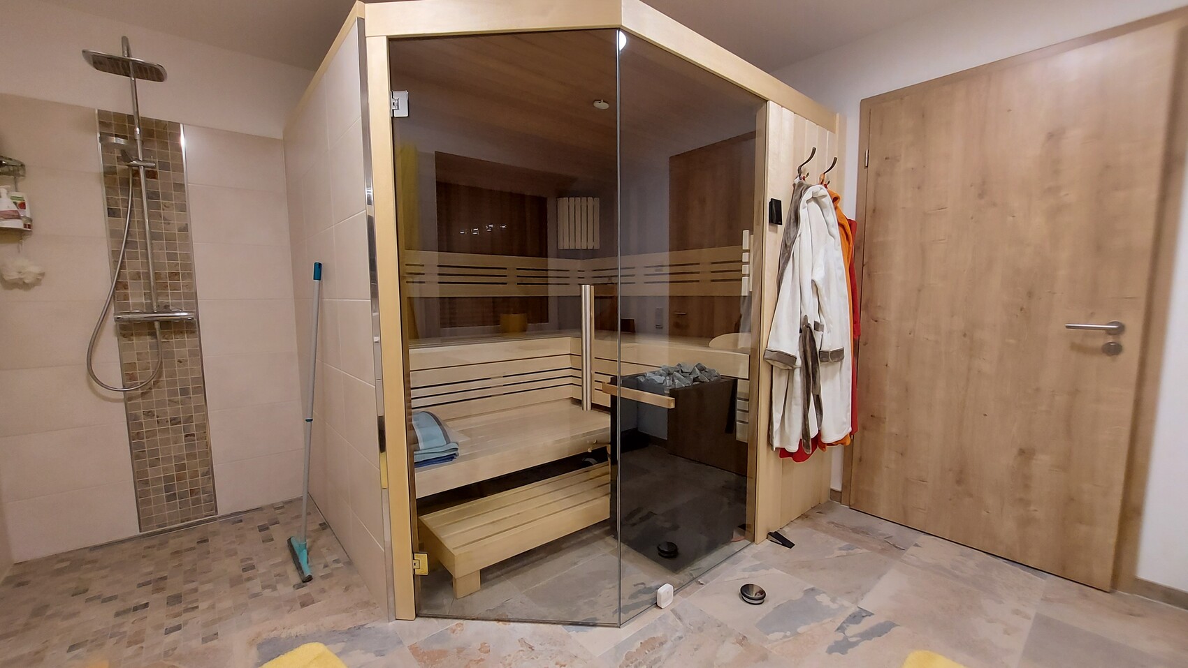 Sauna mit Glasfront und Holzbänken, daneben Dusche und Mantelhaken mit Bademänteln.