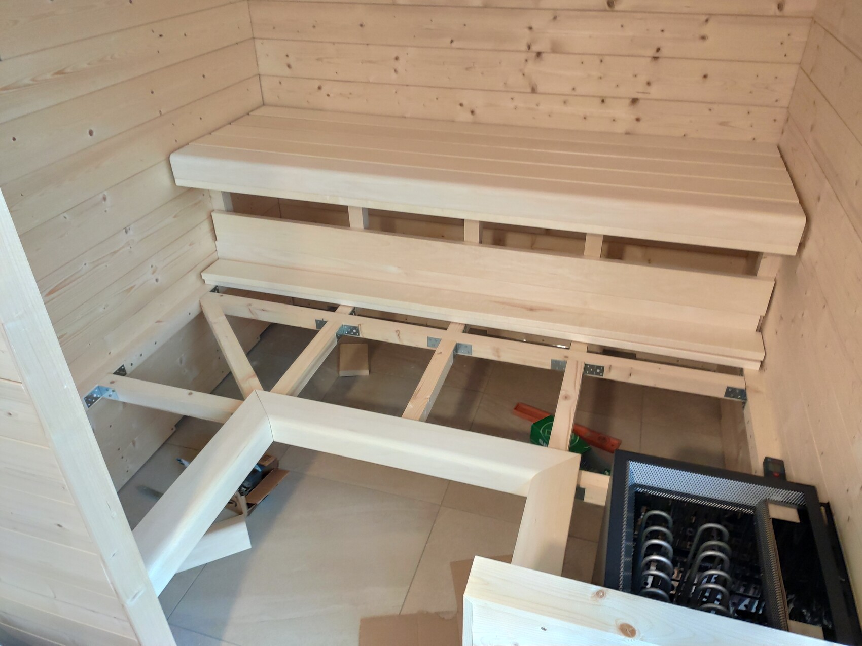 Innenraum einer Holzsauna mit zwei gestaffelten Bänken und Heizgerät am Boden.