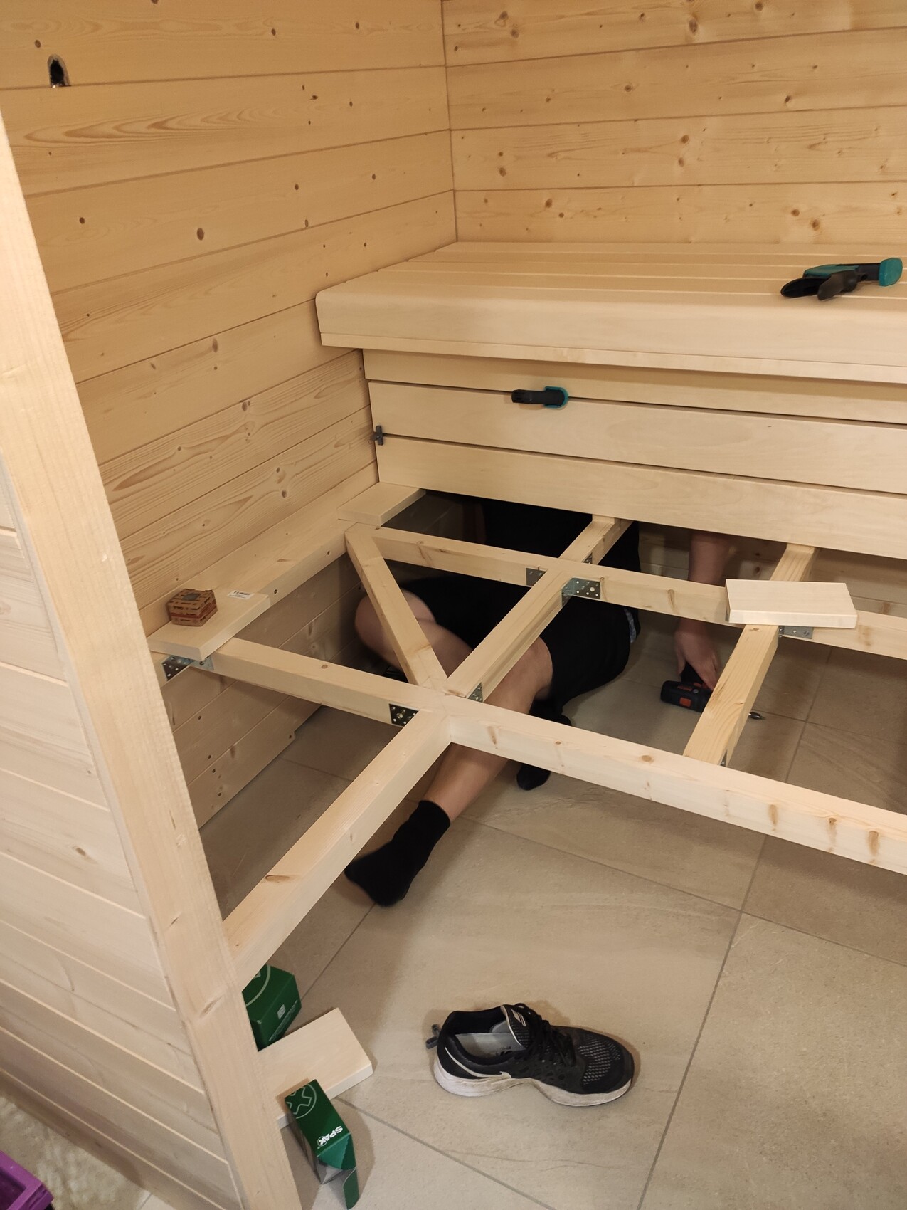 Person arbeitet unter einer Holzrahmenkonstruktion in einem Holzraum; Werkzeuge liegen daneben.