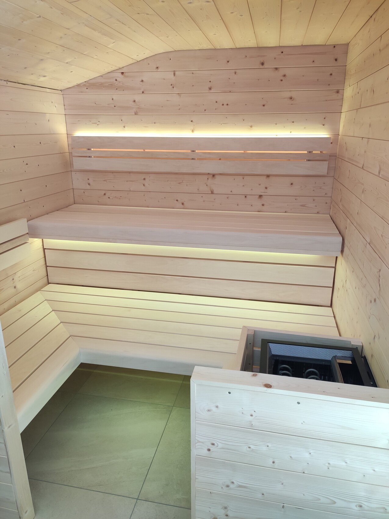 Helle Holzsauna mit mehreren Bänken, Heizofen in der Ecke und warmem Licht.