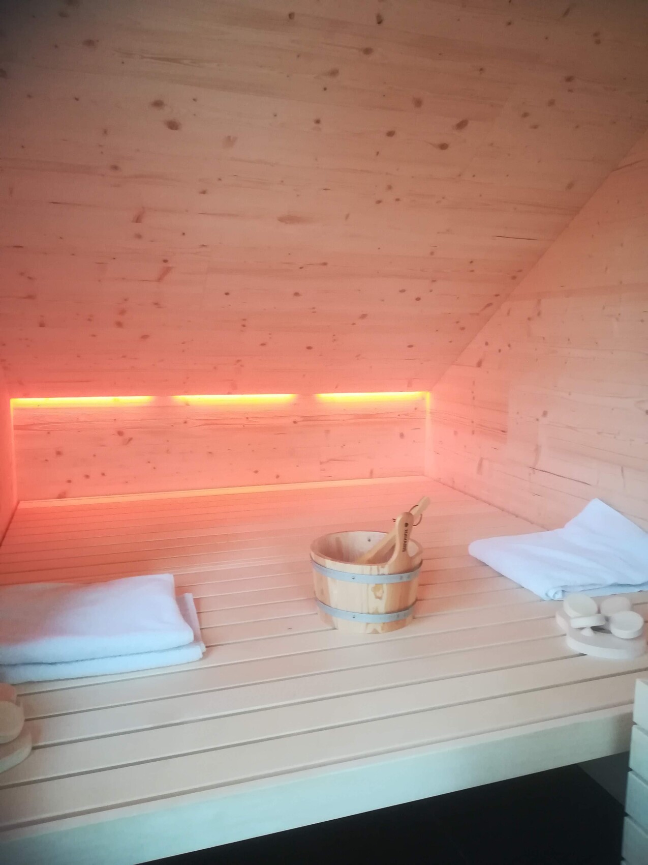Holzsauna mit Bank, Eimer mit Schöpfkelle und Handtücher, warmes orange LED-Licht.