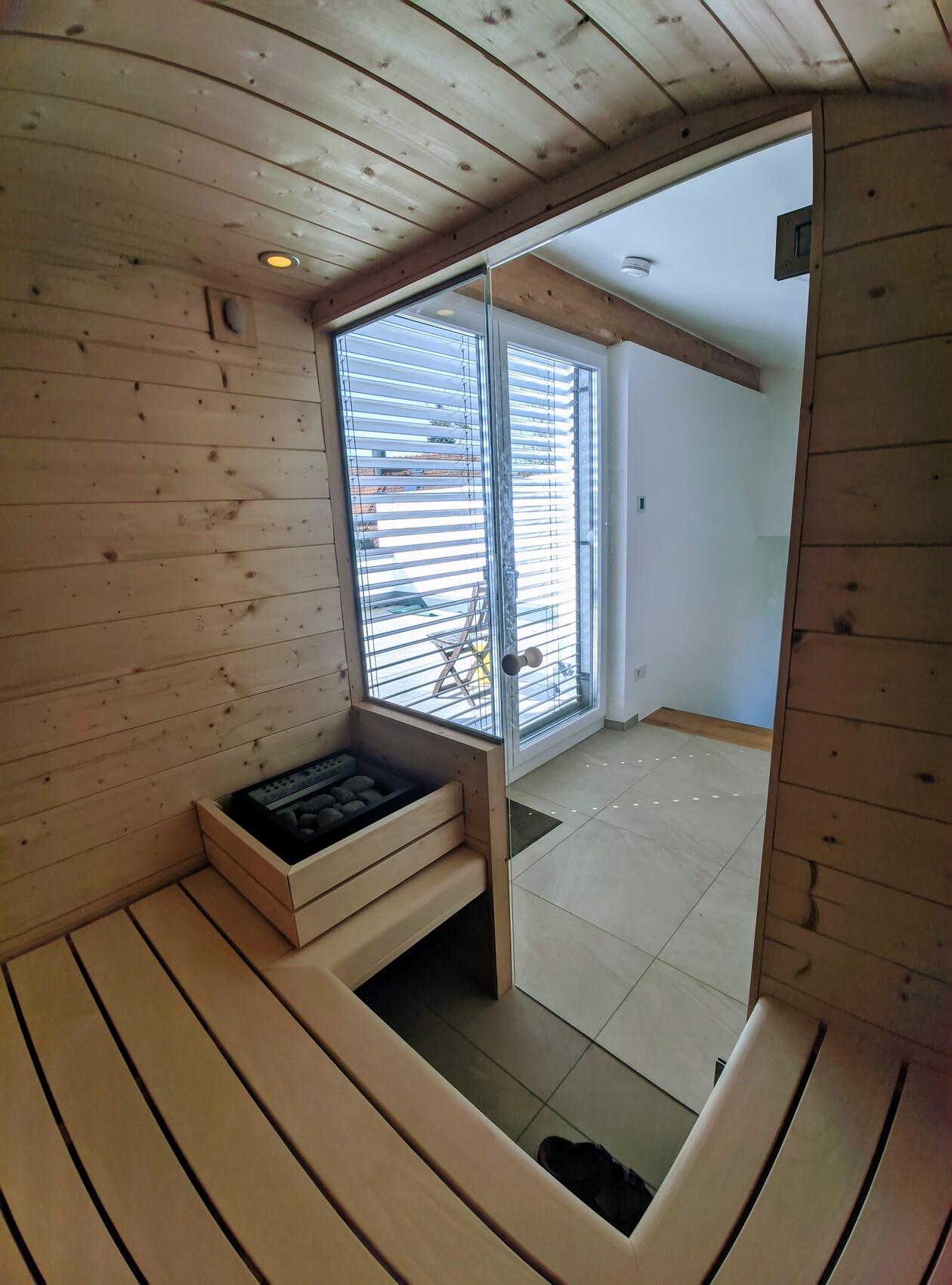 Holzsauna mit gebogenen Bänken, Saunaofen und Glas-Tür zum Außenbereich mit Jalousien.