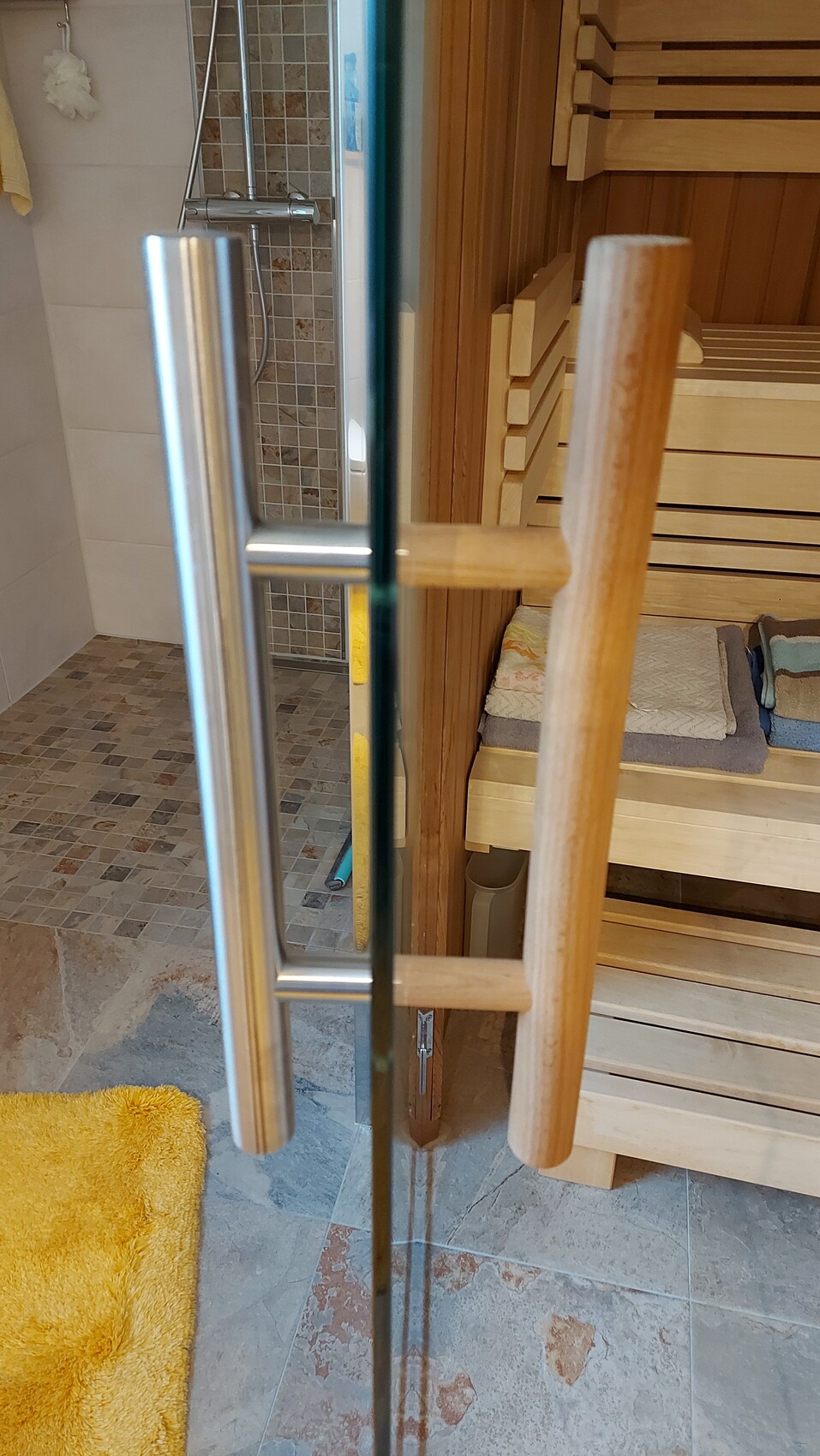 Glastür zum Saunaraum; Holzbänke, Handtücher und gelbes Badetuch im Vordergrund.