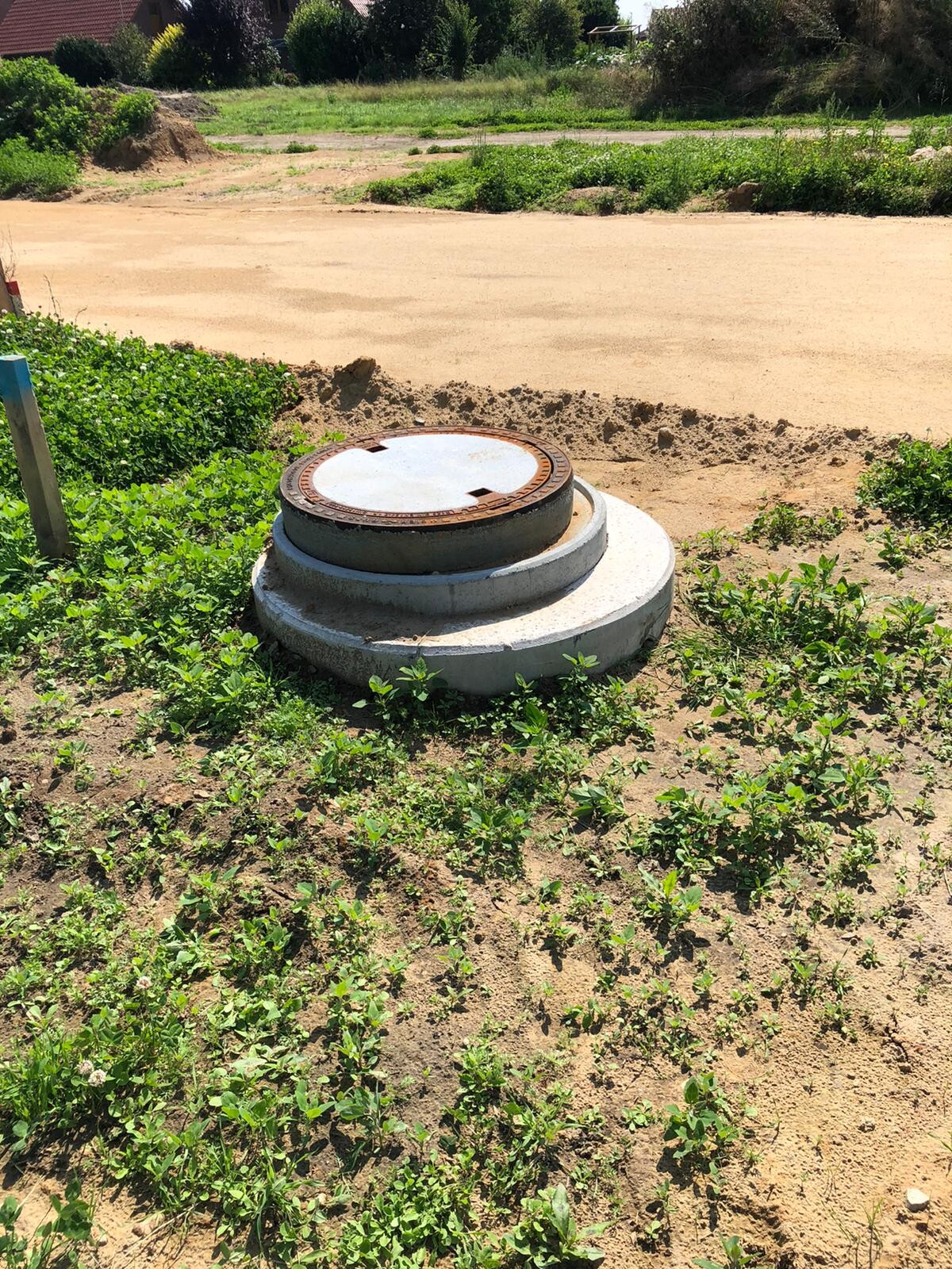 Rostiger Metalldeckel auf grauer Betonplatte in sandigem Gelände, von Gras umgeben.