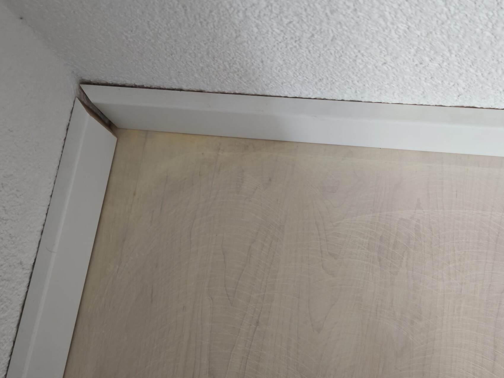 Innenraumecke: Weißer Türrahmen, kleiner Spalt an der Ecke, beige Boden mit Maserung.