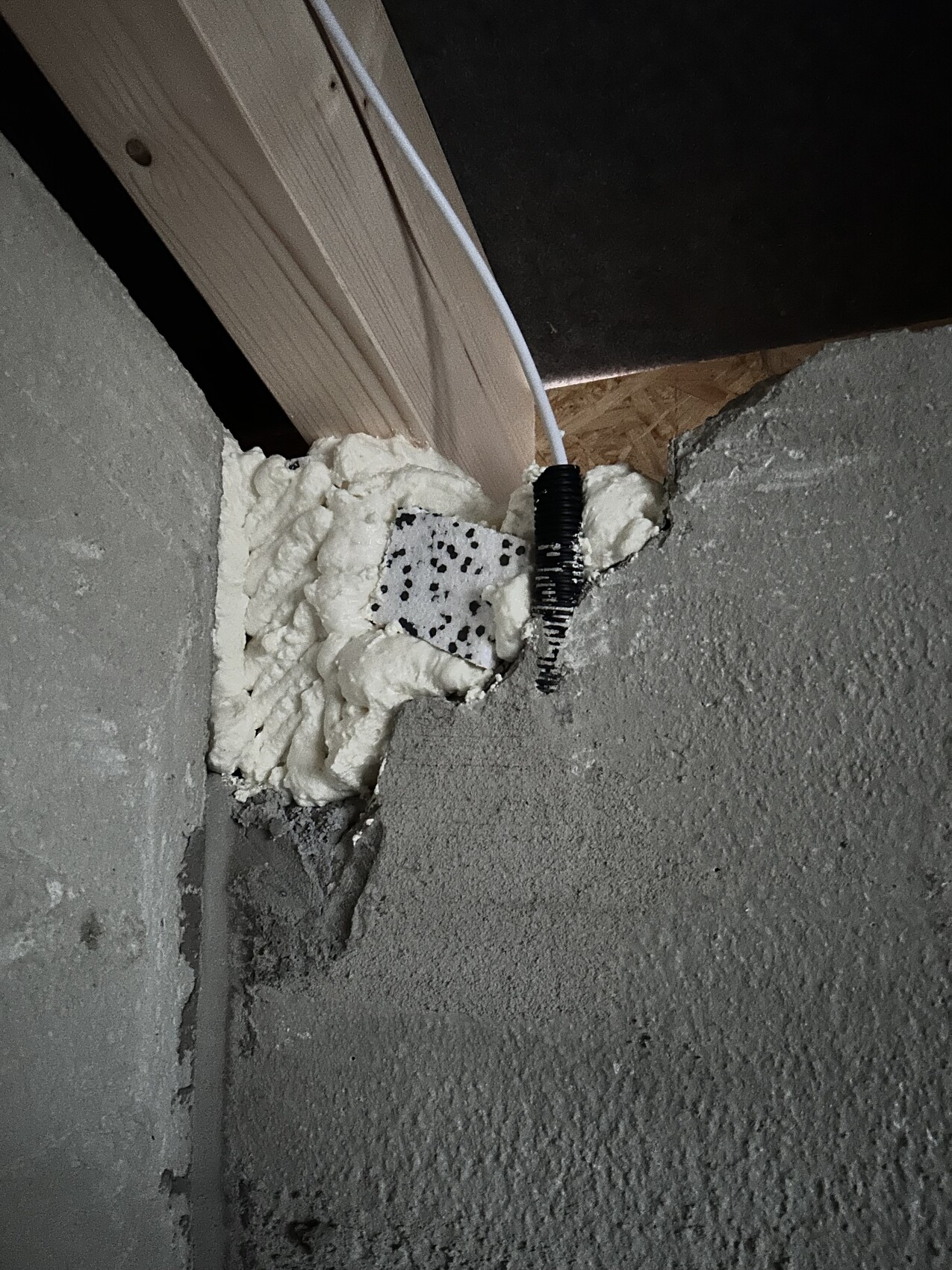 Weißer Dichtschaum füllt Spalte an Betonwand; Kabeldurchführung neben Holzrahmen sichtbar.