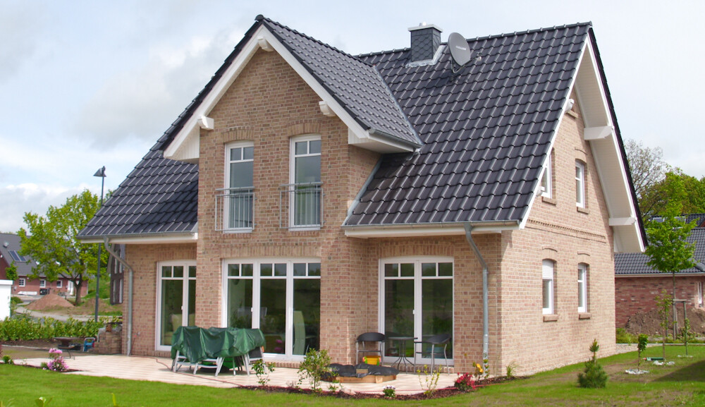 Zweistöckiges Backsteinhaus mit dunklem Ziegeldach, großen Fenstern und einer Terrasse im Garten.