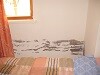 Kleines Schlafzimmer mit Bett, Fenster und orangen Vorhängen, Wand teils beschädigt.
