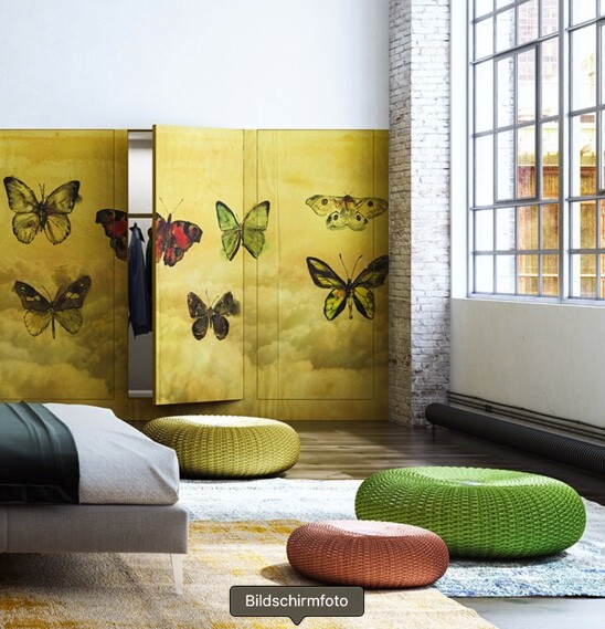 Schlafzimmer mit gelbem Paravent mit Schmetterlingen, Bett links, bunte Poufs, großes Fenster.