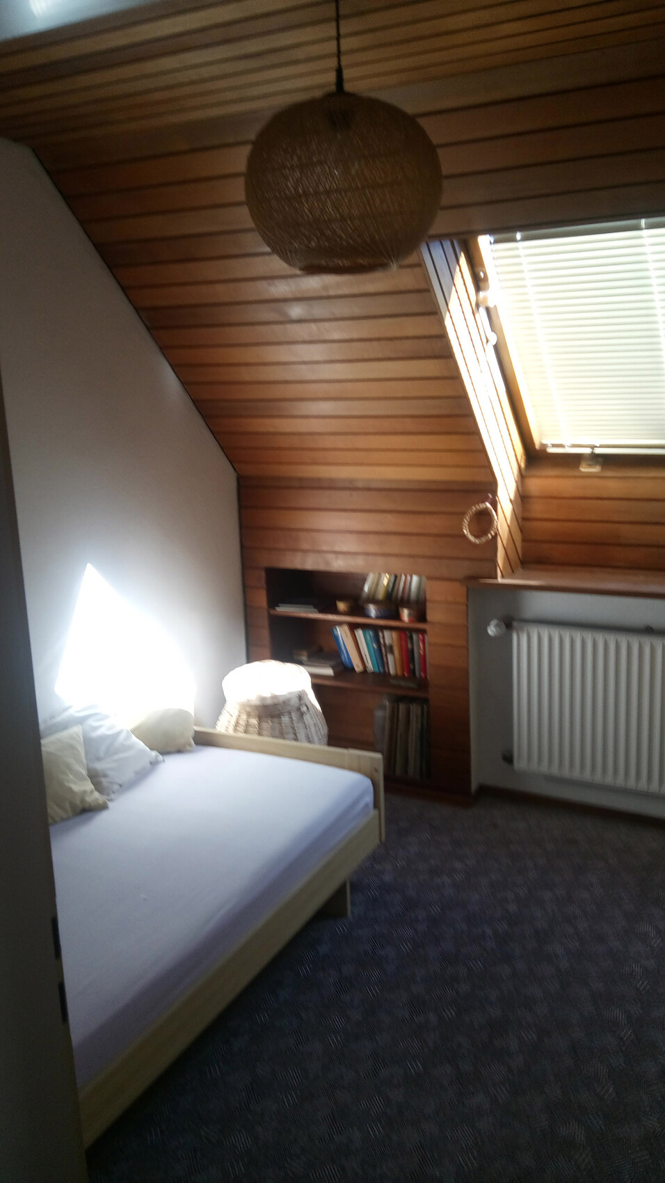 Schlafzimmer mit Holzwandverkleidung, Bett, Bücherregal, runder Lampenschirm, Dachfenster und Heizkörper