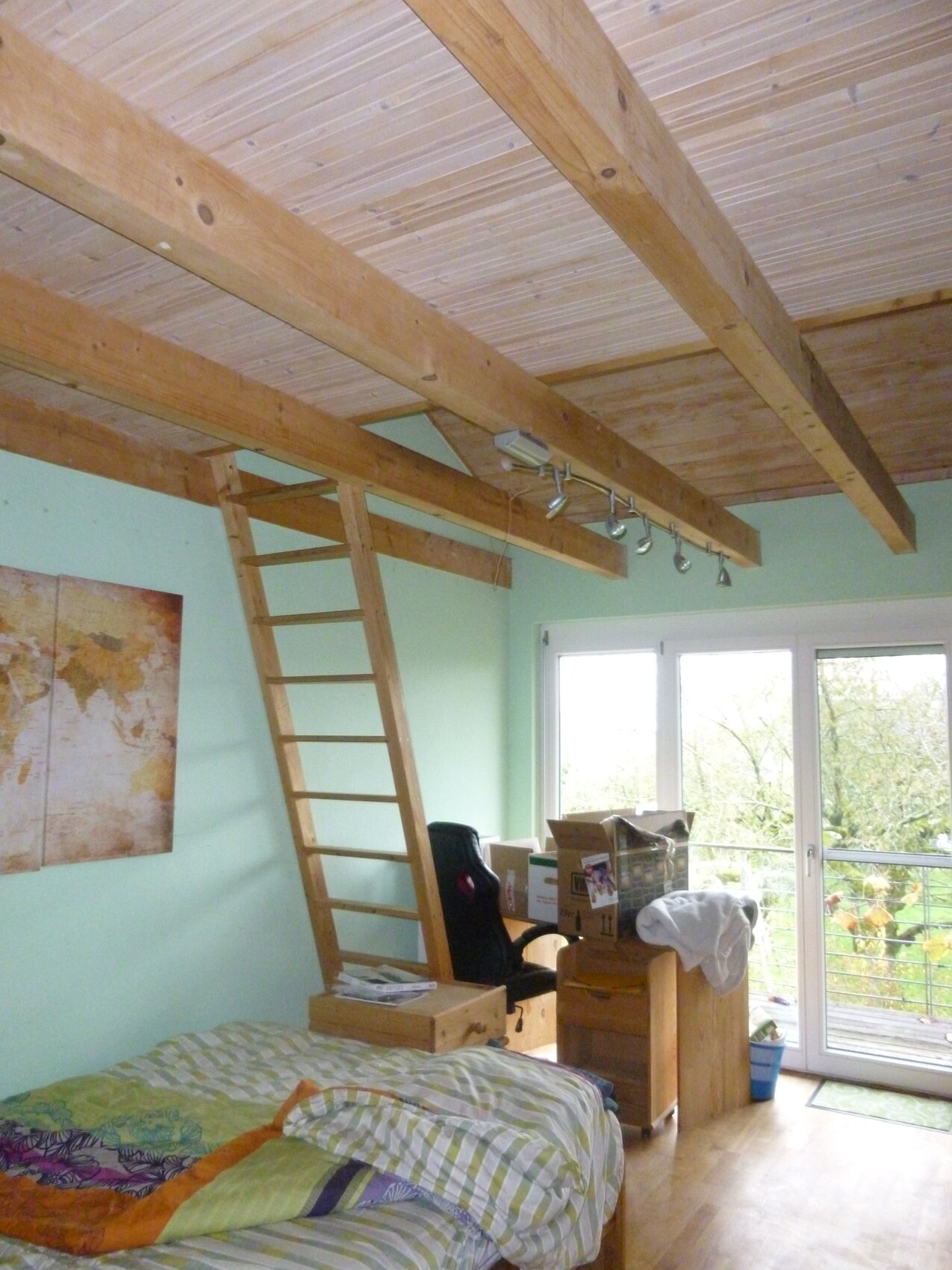 Schlafzimmer mit Holzbalkendecke, Leiter zum Loft, Arbeitsbereich mit Kartons, Bett nah Fenster.