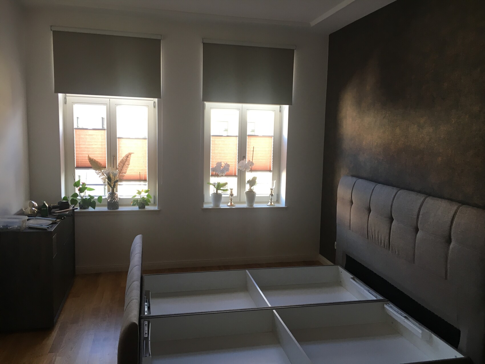 Schlafzimmer mit grauem Polsterbett, ausgezogenen Schubladen, zwei Fenster mit Jalousien, Pflanzen.