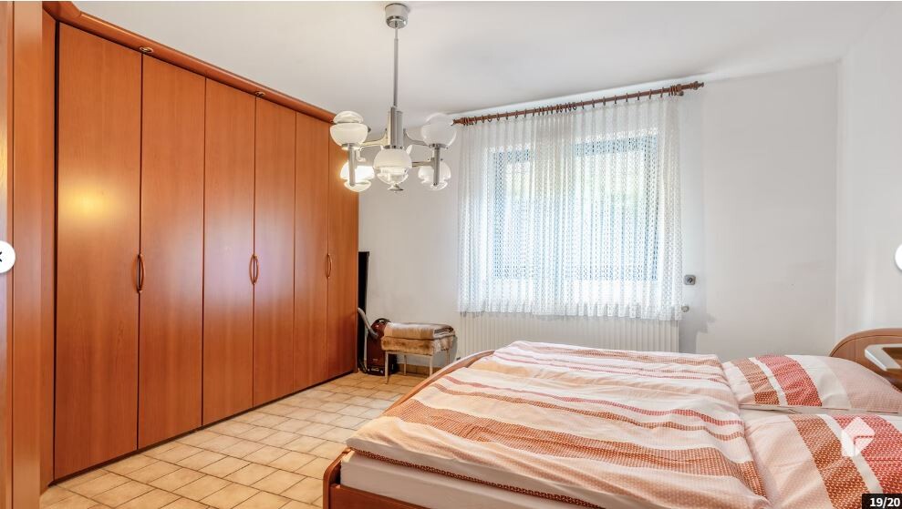Schlafzimmer mit massiver Holzschrankwand, Bett mit gestreifter Decke, Fenster mit Vorhängen.