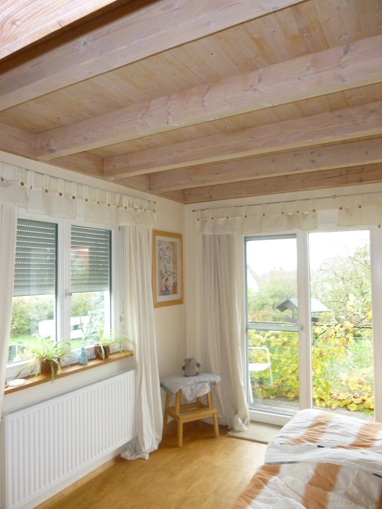 Schlafzimmer mit Holzbalkendecke, großen Fenstern und Balkonzugang, Vorhängen und Pflanzenecke.