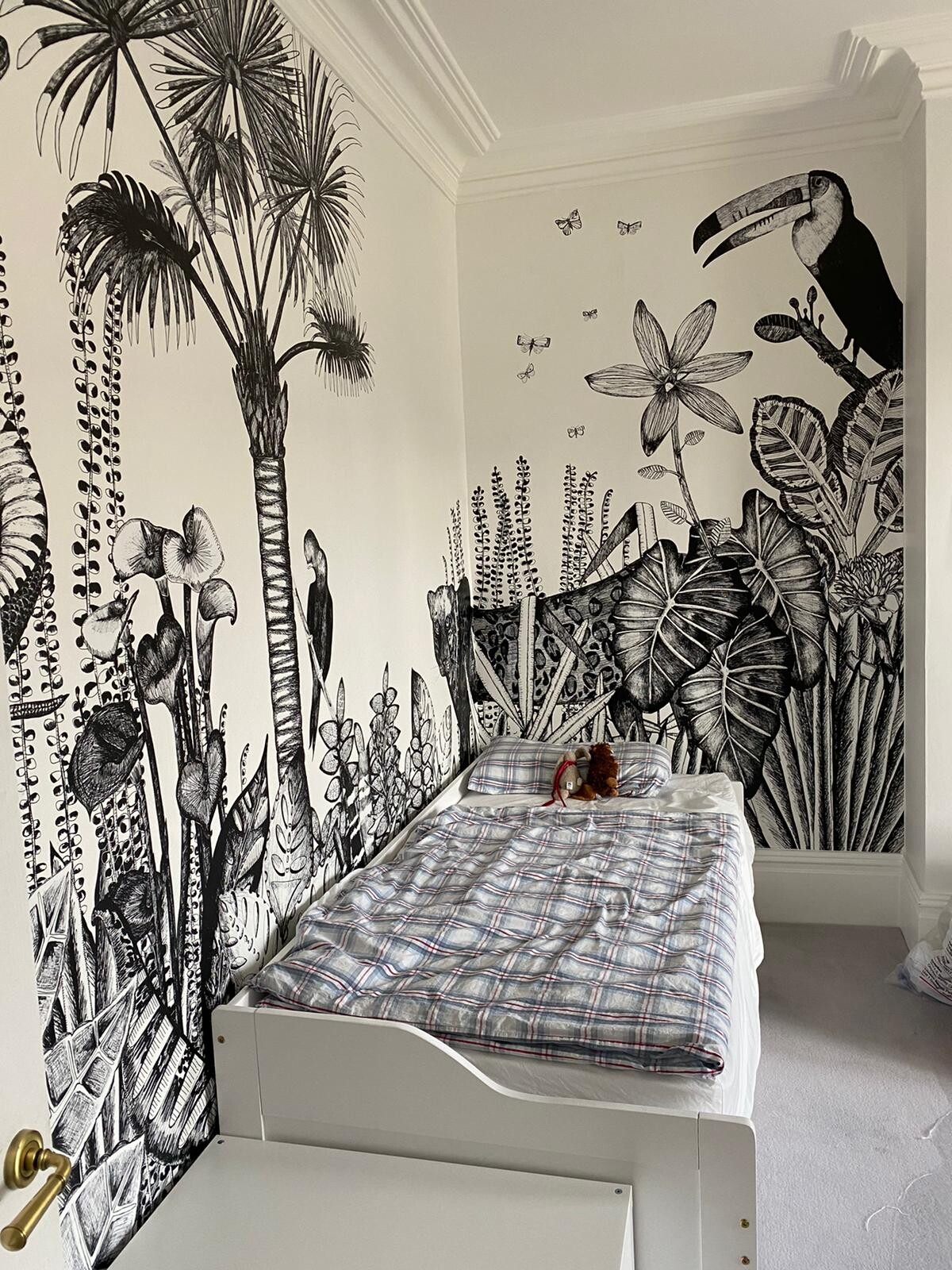 Schlafzimmer mit schwarz-weißer Dschungel-Wandbemalung, Palmen, Toucan, Bett mit Plaid-Decke.