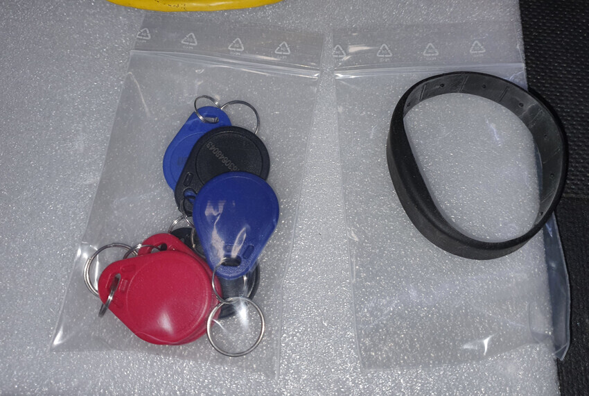 Mehrere runde Schlüsselanhänger in Beuteln (rot, blau, schwarz) neben einem schwarzen Armband.