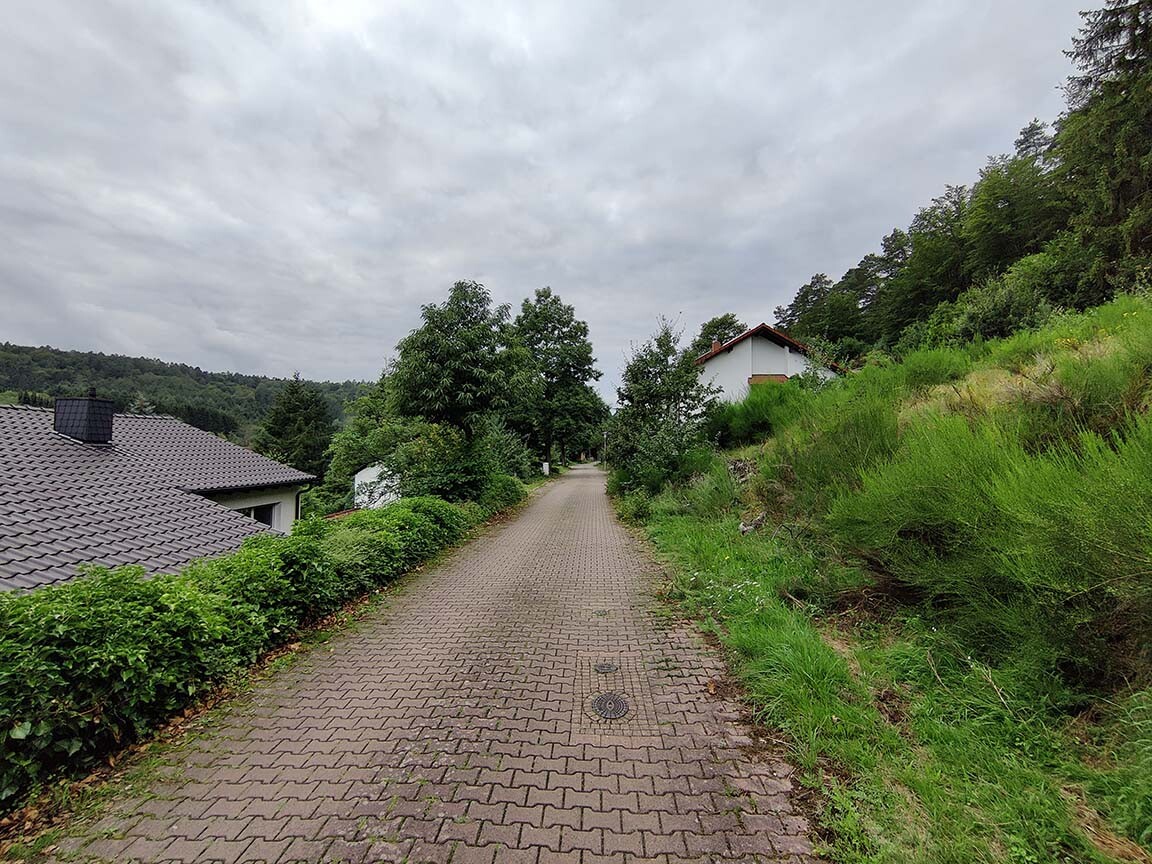Gepflasterter Weg durch grüne Hügellandschaft, Häuser links und rechts, wolkiger Himmel