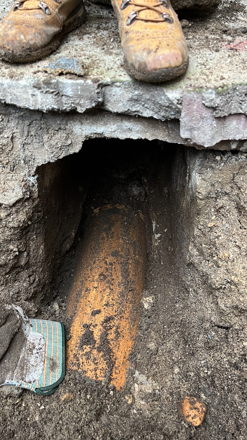 Arbeitsstiefel stehen an der Kante eines Erdlochs; orangebrauner Holzbalken liegt im Loch.