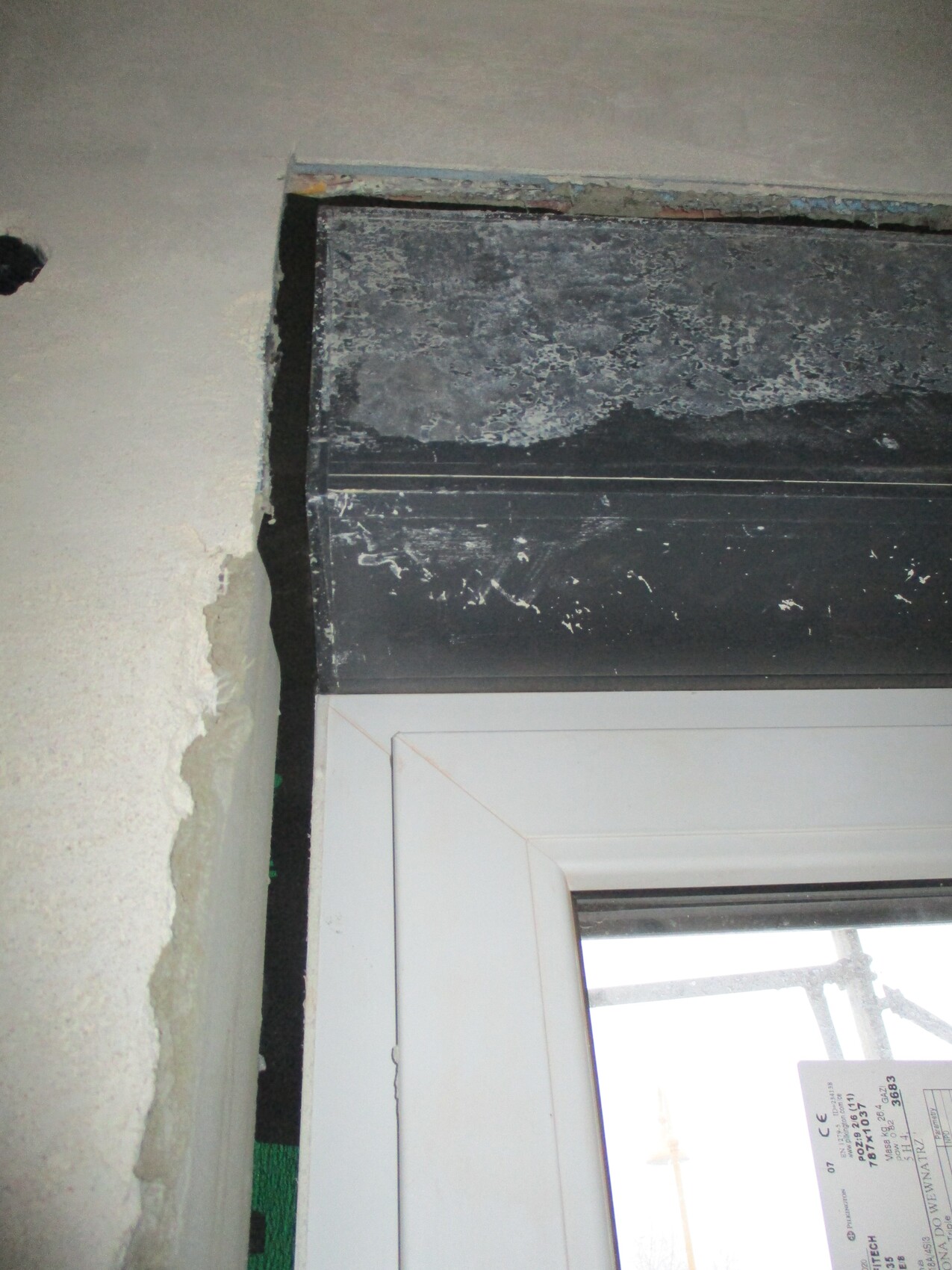 Rohbau: Weißer Fensterrahmen unter schwarzer, verschmutzter Rollladenführung; Putz bröckelt.
