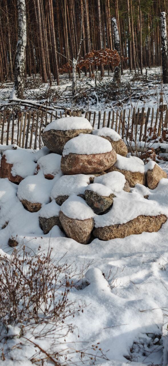 Verschneite Steinpyramide aus runden Steinen im Wald, hinter Holzzaun.