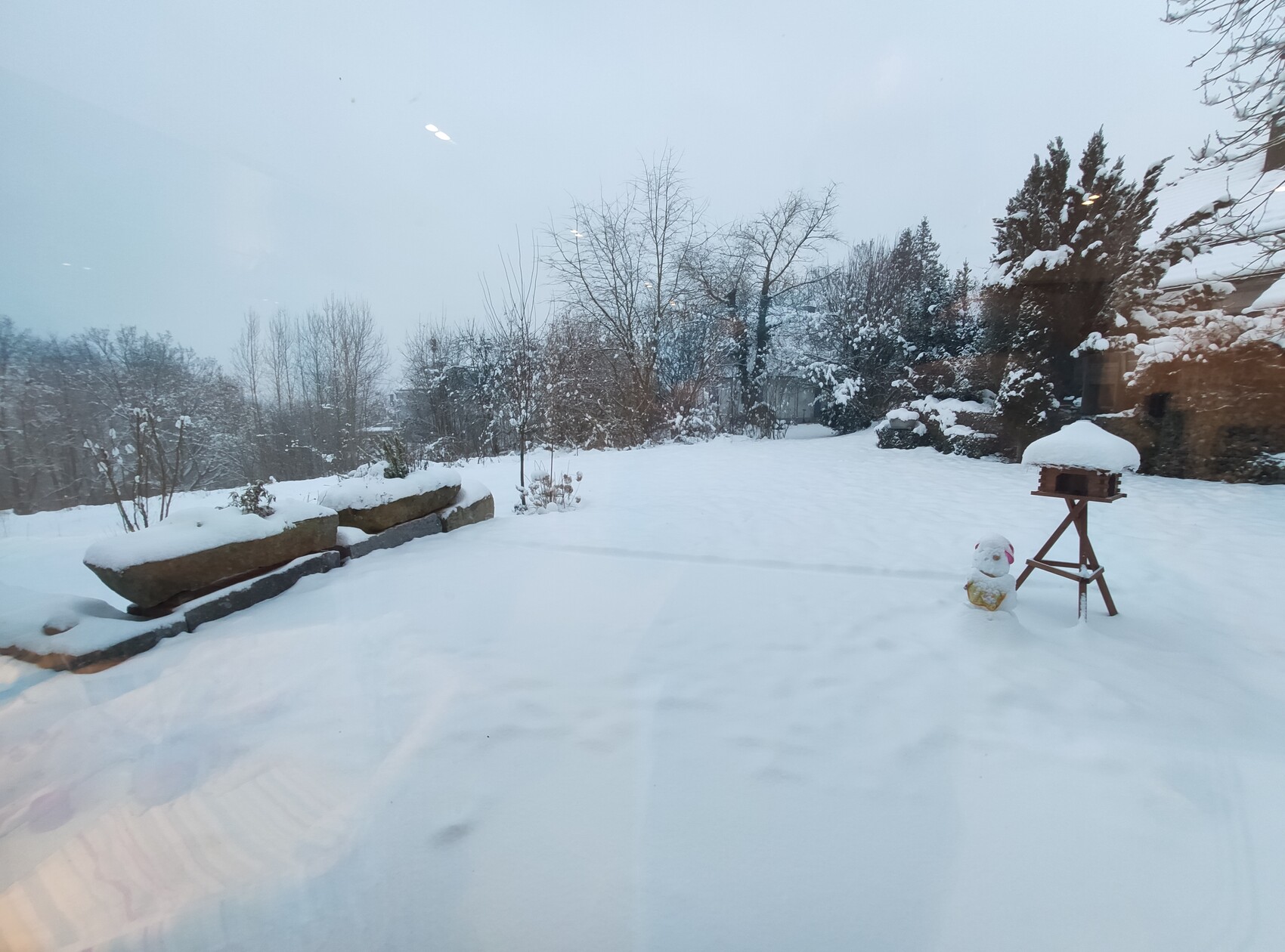 Garten im Winter mit schneebedecktem Rasen, Vogelhaus auf Stativ und Schneemann im Vordergrund