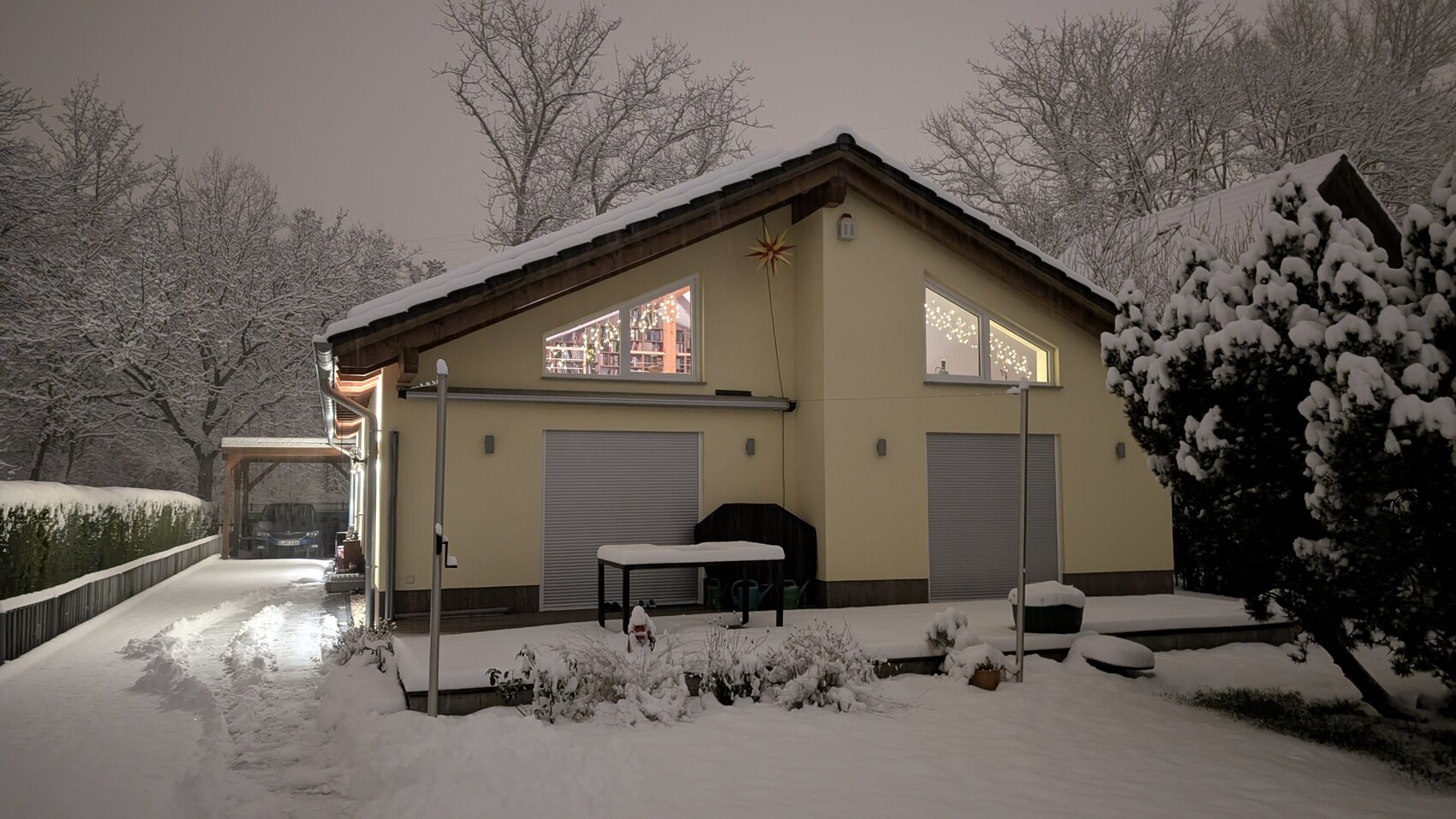Zweistöckiges gelbes Haus mit Schnee auf dem Dach, Fenster mit Lichtern, verschneiter Garten.